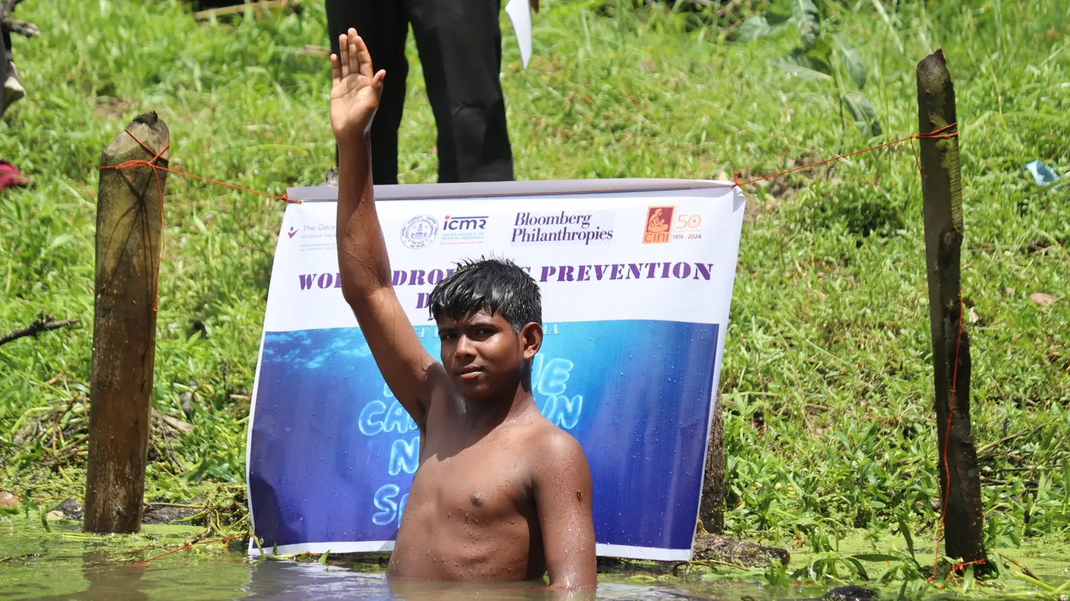 An adolescent raising his hand during a pond-based drowning-prevention activity with partner signage behind him
